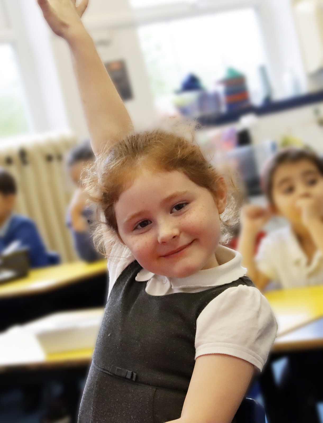 A smiling young girl pupil holds her hand aloft ready to be picked to answer the question from the teacher