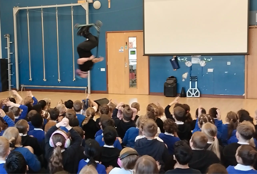 Man somersaulting on a scooter in front of a school assembly